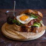 A bacon and egg sandwich on wooden board with a pot of relish