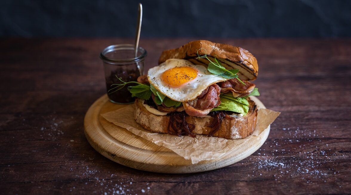 A bacon and egg sandwich on wooden board with a pot of relish