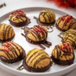 Nine chocolate decorated macaroons on a white round plate and pistachios and red flower in the back