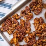 A corner of a rectangle tray with brown nuts, a wooden spoon full of cinnamon powder on green striped cloth, and a tray of cinnamon sticks