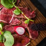 pieces of thin sliced meat on wooden board topped with herbs , radish and blackberry.