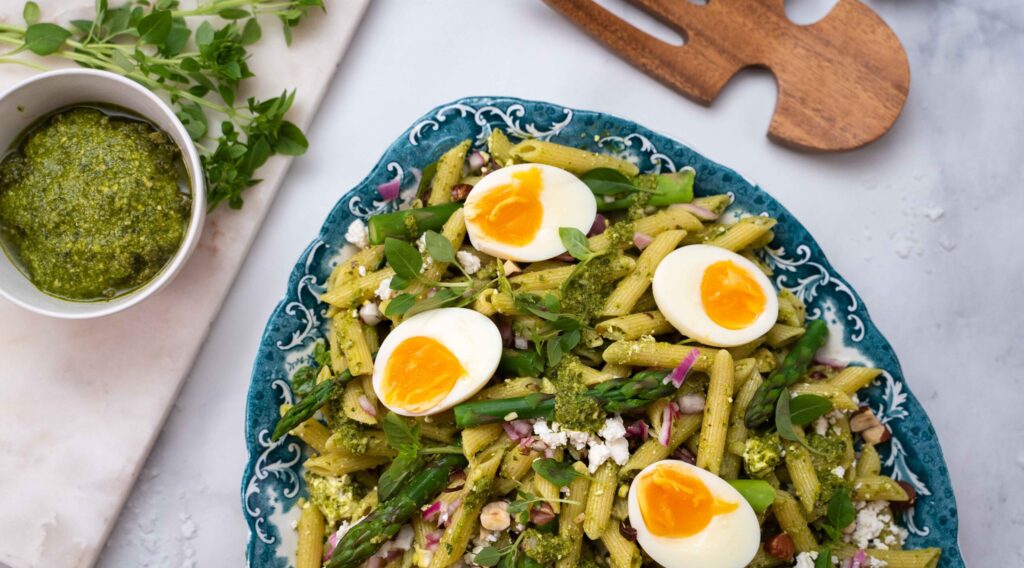 A part of large platter full of penne pasta and asparagus salad in green dressing topped with four halves of hard boiled egg. Salad servers, a pot of pesto sauce and herbs.