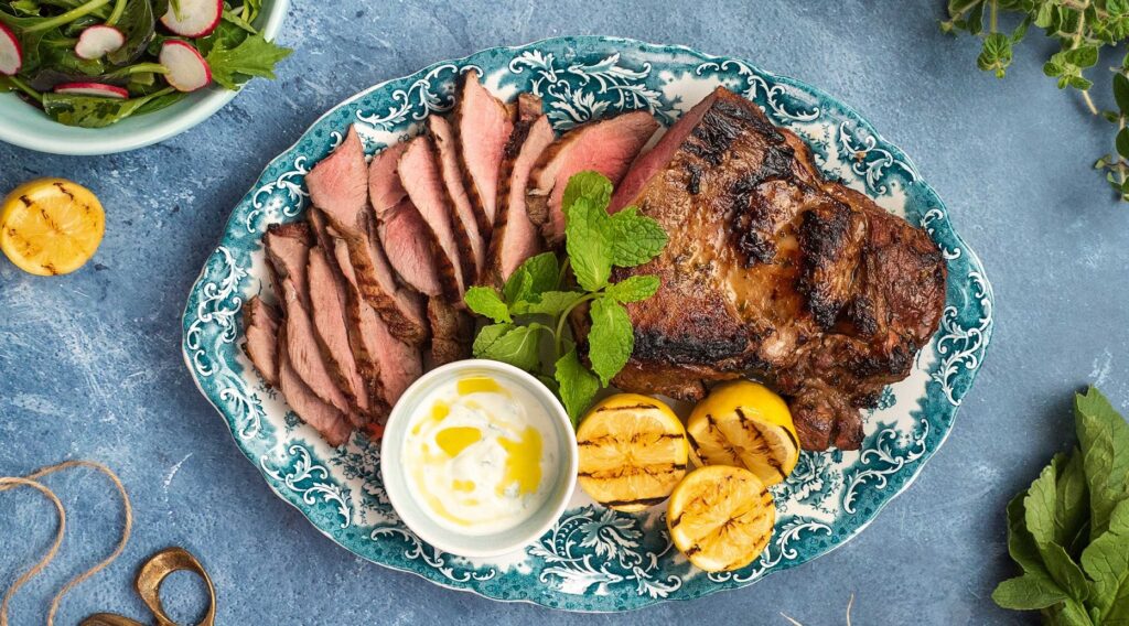 A platter of sliced meat, a pot of white sauce and grilled halves of lemon on blue bench top , a bowl of salad, green herbs around it.