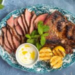 A platter of sliced meat, a pot of white sauce and grilled halves of lemon on blue bench top , a bowl of salad, green herbs around it.