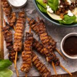 Several grilled meat skewers, a bowl of greenish salad, small bowls of dark sauce and salt, basil on blue background