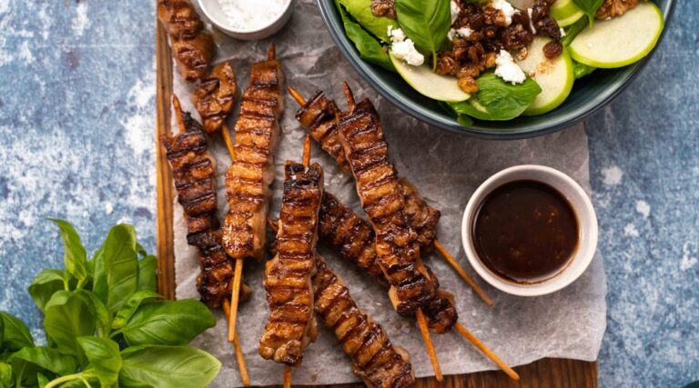 Several grilled meat skewers, a bowl of greenish salad, small bowls of dark sauce and salt, basil on blue background