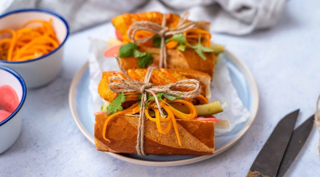 Two pieces of baguette filled with broen, orange and green food each tied with a string on blue plate, small pots of carrots and radish, a pair pf scissors.