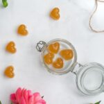 top view of an open jar with four hart shaped sweets inside, more sweets outside, a flower, pot of honey, string and scissors on white bench top
