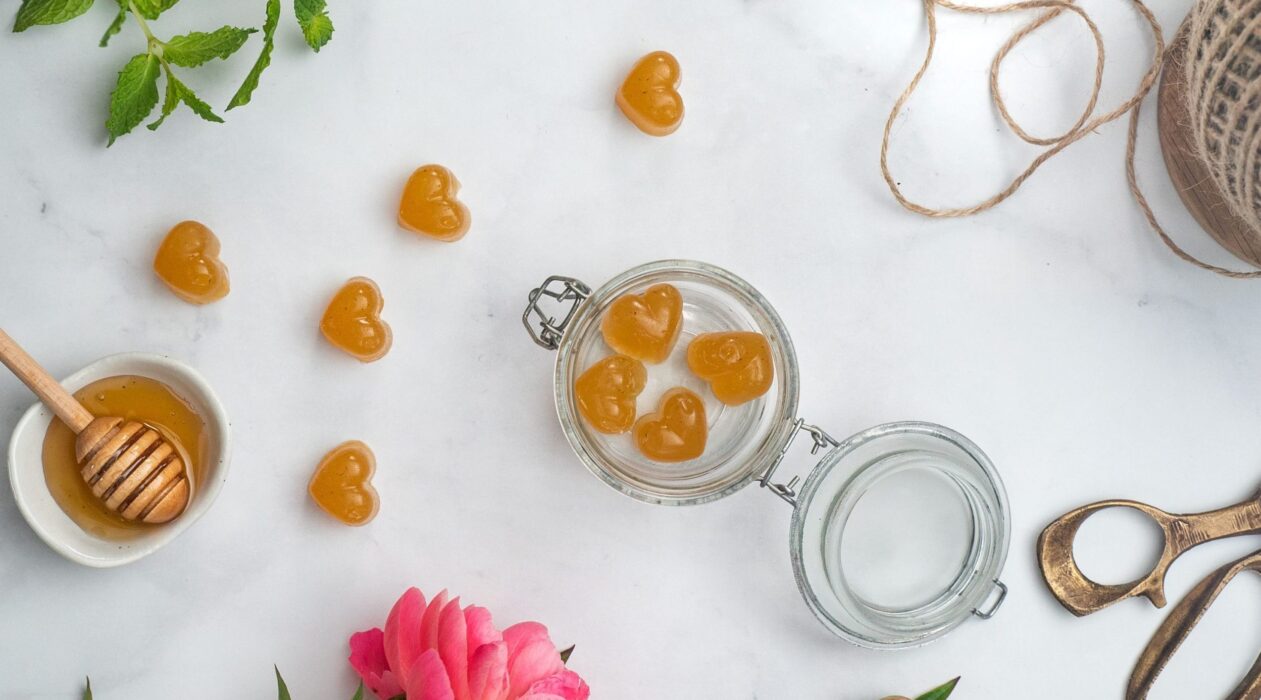 top view of an open jar with four hart shaped sweets inside, more sweets outside, a flower, pot of honey, string and scissors on white bench top
