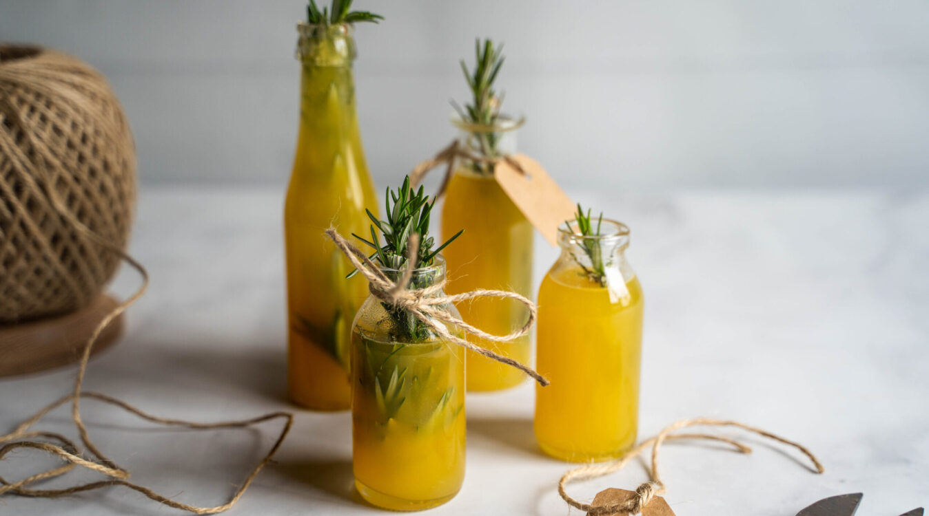 Four small bottles full of yellow liquid with rosemary sprigs, strings on white bench top.