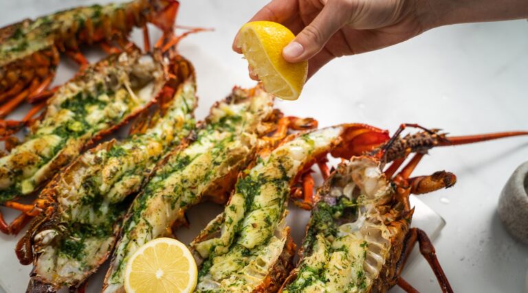 Six cooked crayfish halves with herb on top, a hand squeezing a lemon half and a cut lemon on food.