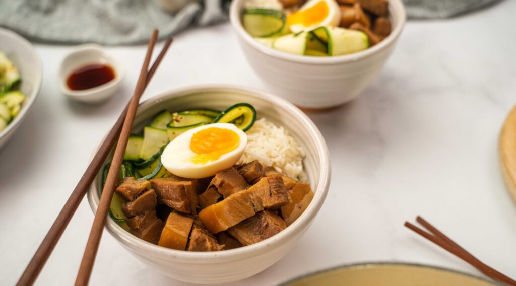 Two bowls of cooked meat , courgettes and boiled egg on rice with chopsticks on a white table with cloth, pot of dark sauce