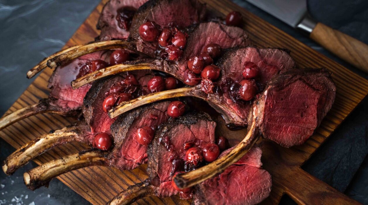 Ten cutlets of pink cooked meat on wooden board topped with red cherries and a knife