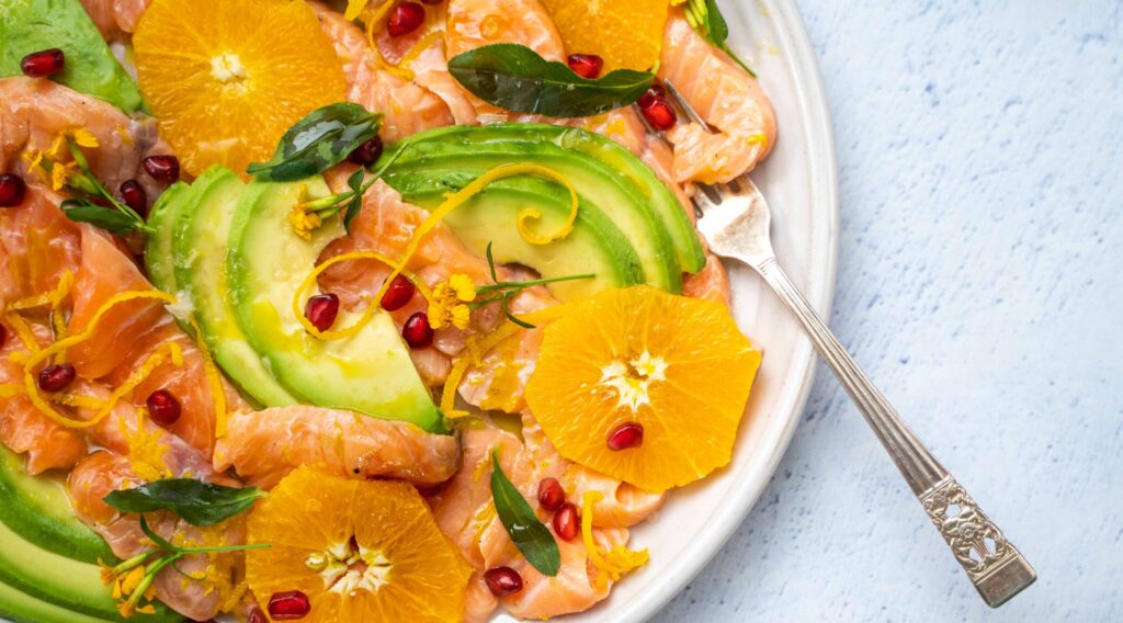 Orange, avocado and salmon slices on a plate topped with red berry, citrus zest and herb with a fork on pale blue table top