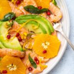 Orange, avocado and salmon slices on a plate topped with red berry, citrus zest and herb with a fork on pale blue table top