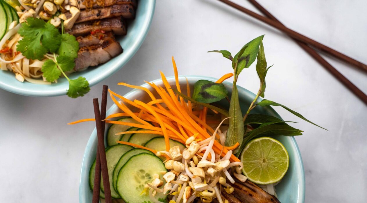 Two bowls full of carrot, cucumber, mint leaves, bean sprouts, pork pieces with chopsticks on white marble.