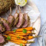 Nut crusted frenched lamb rack pieces on a plate with carrots and pot of white sauce