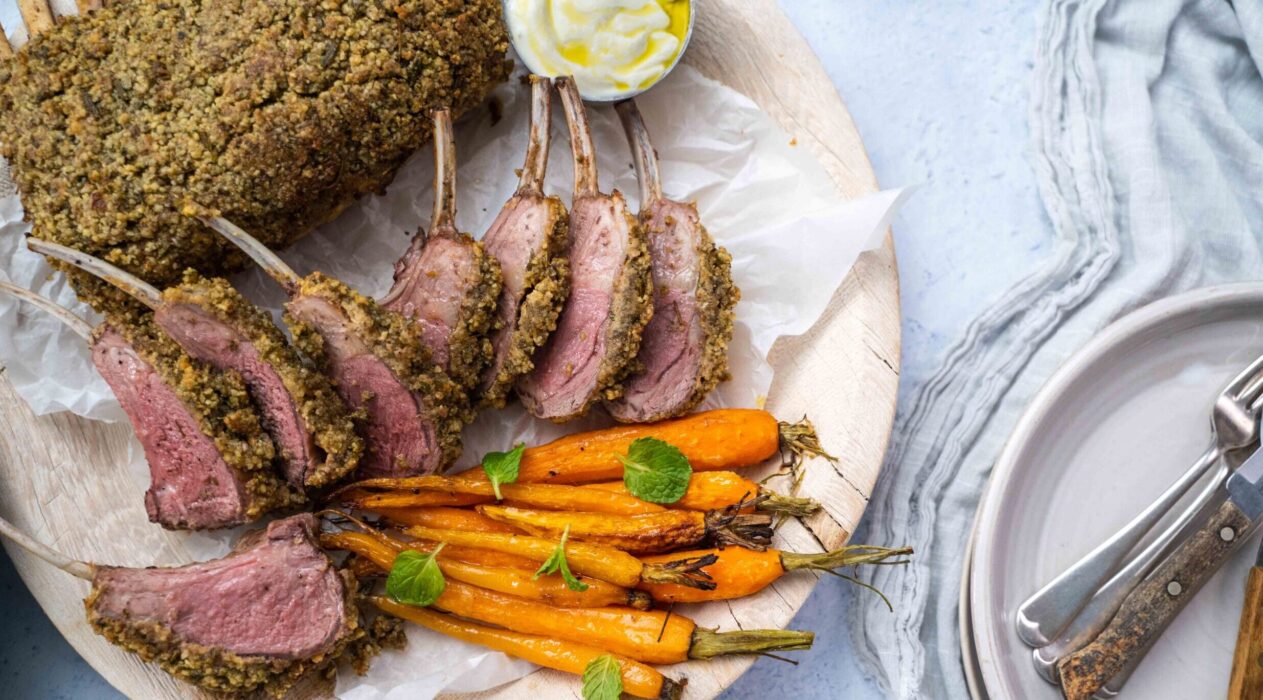 Nut crusted frenched lamb rack pieces on a plate with carrots and pot of white sauce