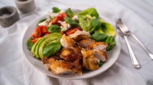A white bowl of salad consists of chicken pieces, avocado, tomatoes, lettuce with cutlery on white cloth, pots of salt and pepper.