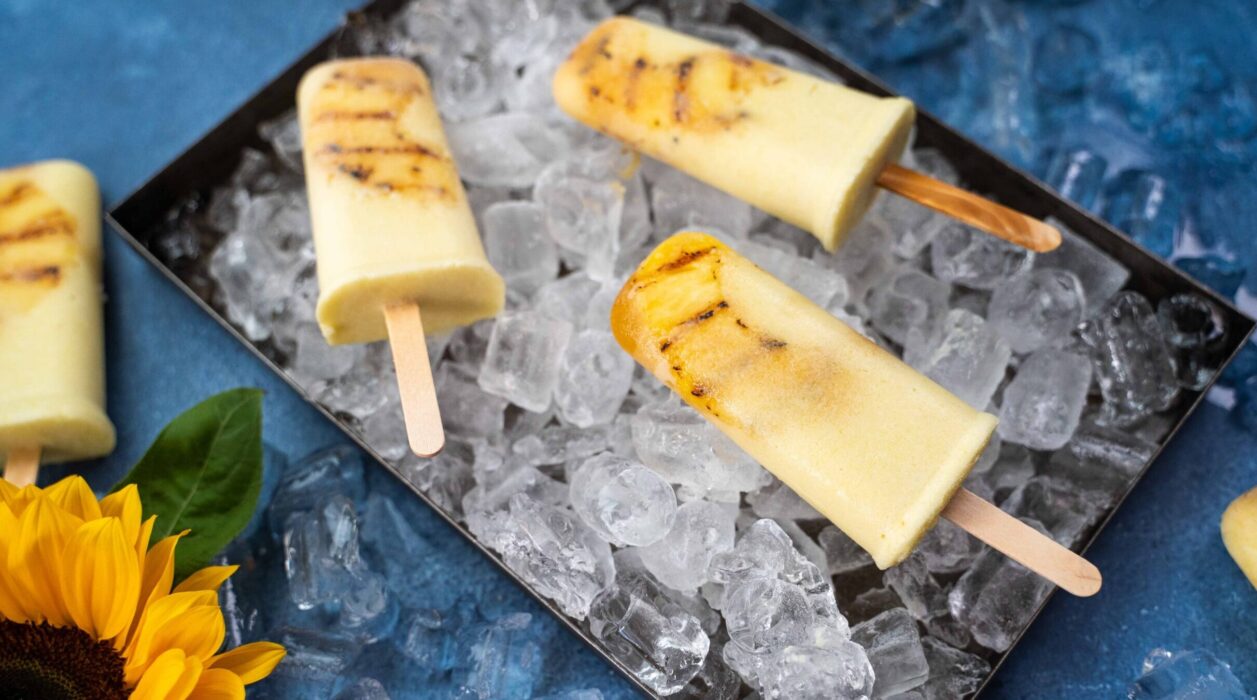 Four creamy coloured popsicles on bed of ice cubes in a tray on blue background with a sunflower.