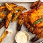 A roast whole chicken topped with rosemary, wedges and a pot of white sauce on a metal tray, and a knife.