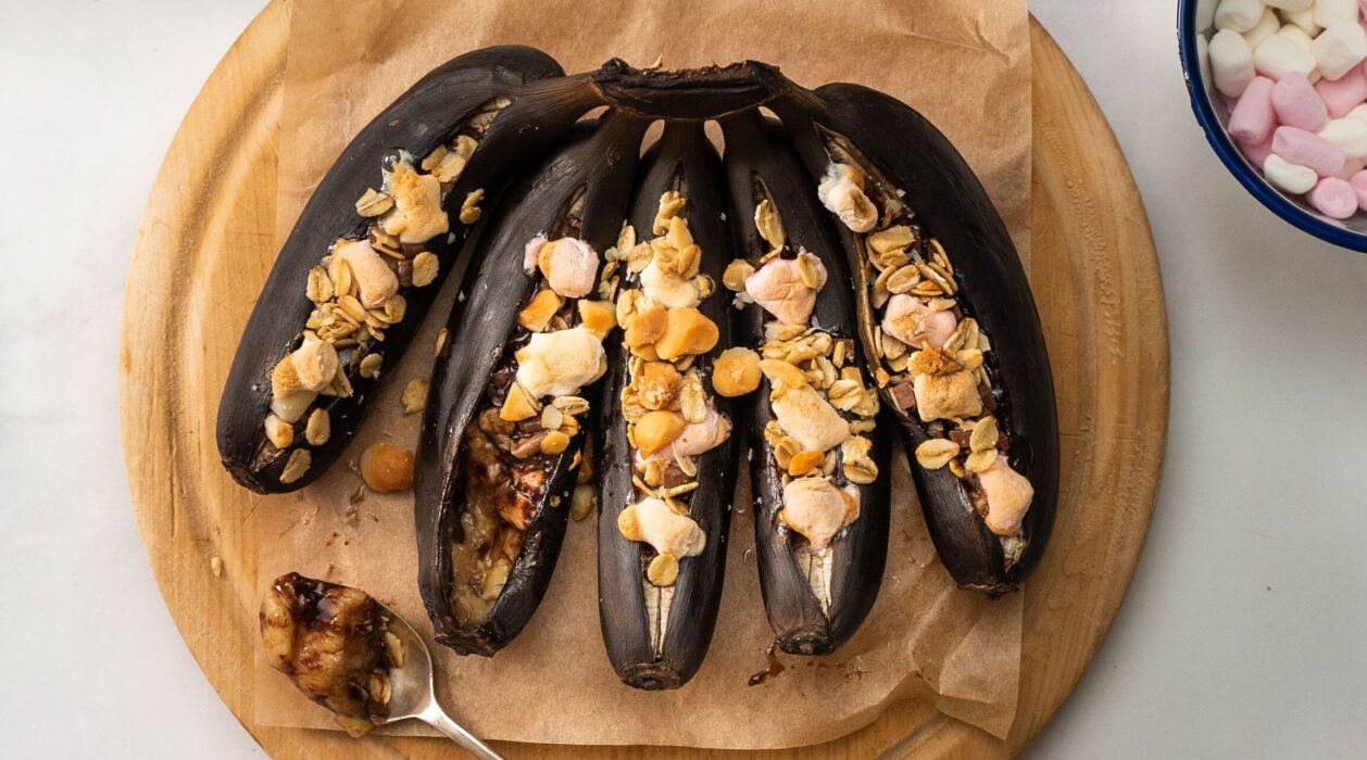 Five blackened bananas topped with pink and brown toppings on wooden board and a bowl of pink and white sweets