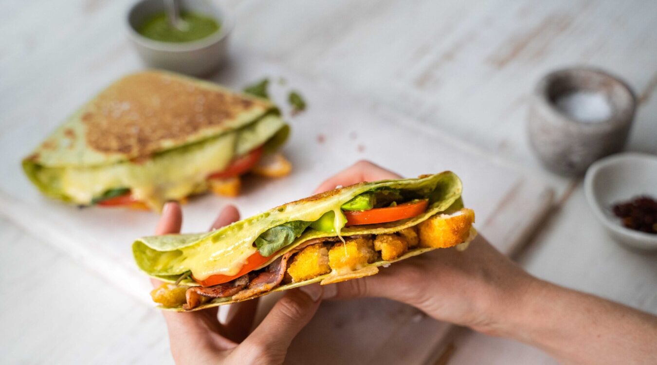 Two hands folding a sandwich made with tortilla, filled with lettuce, avocado, tomato, bacon and crumbed meat. One more sandwich nd a pot of green sauce in the back.