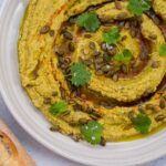 Greenish brown hummus with dark brown spiral pattern with green herb scattered on top on white plate and a bread stick on blue table.