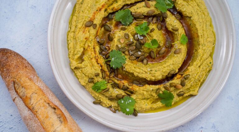 Greenish brown hummus with dark brown spiral pattern with green herb scattered on top on white plate and a bread stick on blue table.