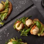 Two open sandwich with creamy coloured balls and greens on black slab, salt flakes scattered around