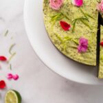 Round green cake with a slice cut topped with flowers on marble, lime halves and more flowers scattered.
