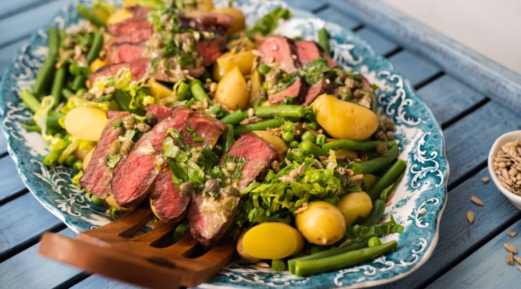 A blue platter of potato, meat and green salad with a wooden salad server on a large blue tray.