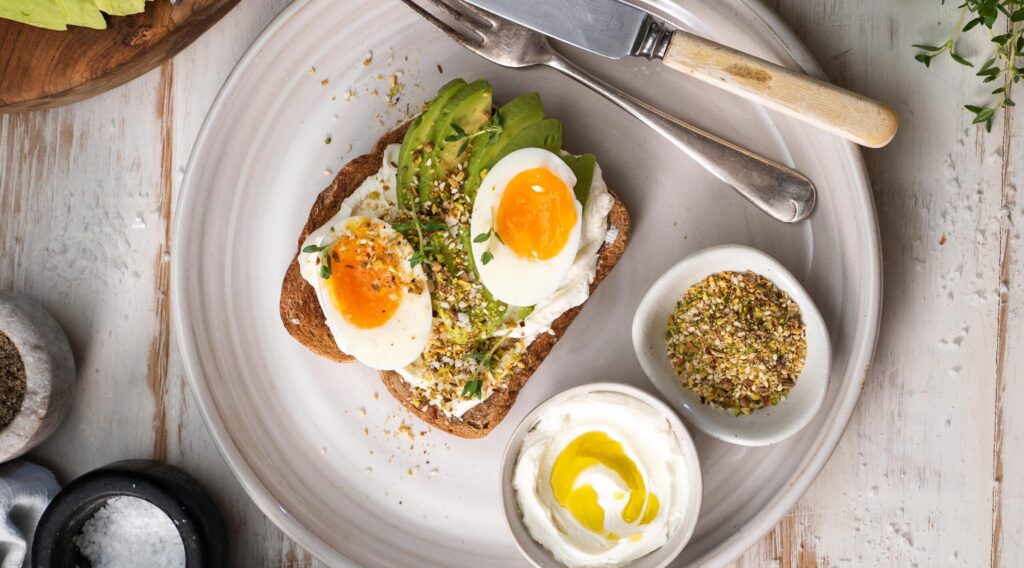 Two halves of an boiled egg, avocado slices and nutty sprinkle on toast on a white round plate with two pots of white cream 7 nutty sprinkles, fork knife on white wooden table.