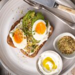 Two halves of an boiled egg, avocado slices and nutty sprinkle on toast on a white round plate with two pots of white cream 7 nutty sprinkles, fork knife on white wooden table.