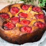 A whole baked cake with red plums on white papered plate, green plant on the right side back