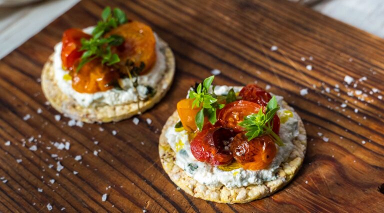 Two round biscuits topped with white cream, baked tomatoes and herbs on wooden board, salt flakes scattered around.