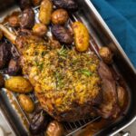 A roasted leg of lamb and potatoes around it in a silver roasting tray, blue cloth, small pots of salt pepper