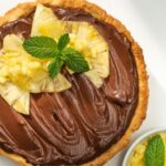 A large tart filled with chocolate cream, topped with pineapple slices and mint leaves on white board with a bowl of cut pineapples and green striped cloth on right side