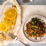 White fish topped with 3 orange slices on a baking paper with a fork, a plate of colourful lentil salad on the right side of it.