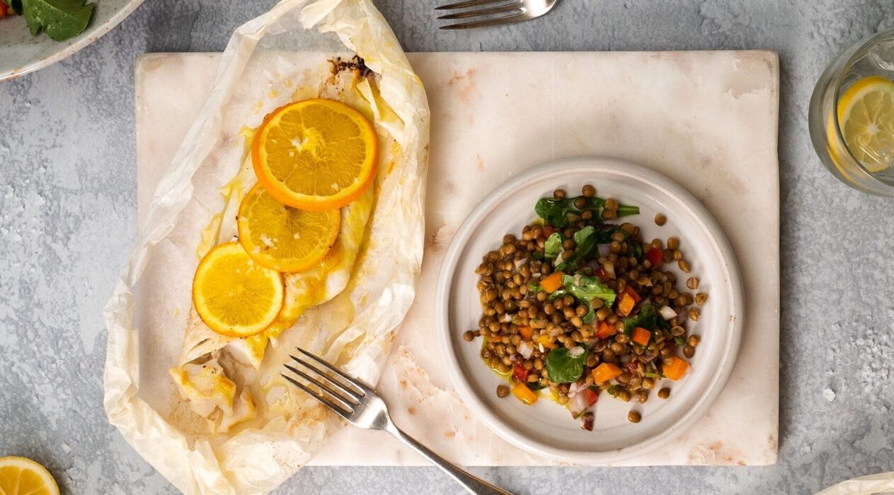 White fish topped with 3 orange slices on a baking paper with a fork, a plate of colourful lentil salad on the right side of it.