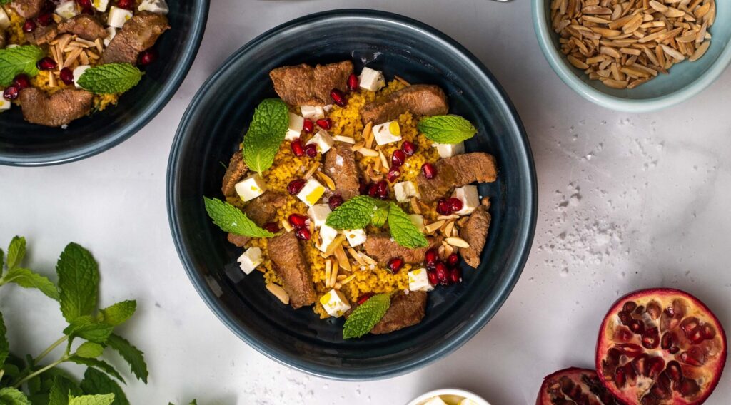 A blue bowl filled with yellow grains topped with brown meat pieces, white cubed cheese, green herb and red seeds, food around it.