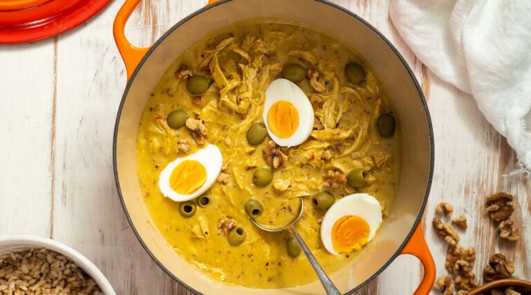 Yellow stew with three boiled egg halves, green olives and shredded chicken in orange coloured caseserole pot with a spoon on white board.