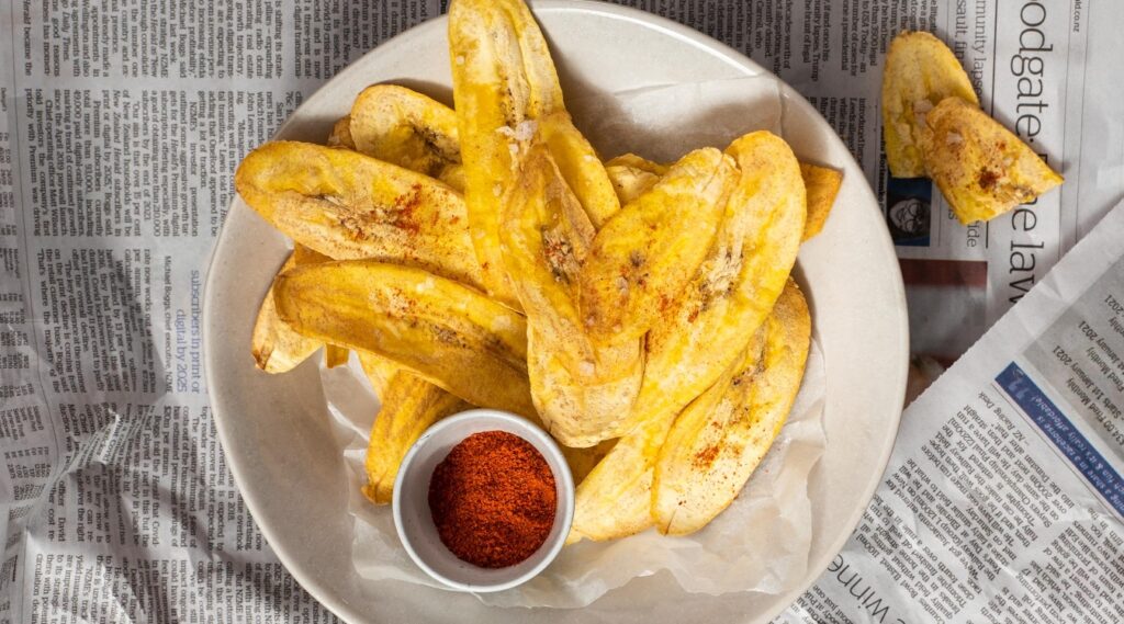 Several yellow banana chips in a shallow white bowl with a small dish of red powder, on newspaper.