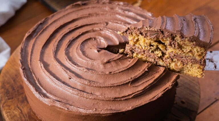 Close-up of a large round chocolate cream cake on a wooden board with a slice lifted up to reveal layers.