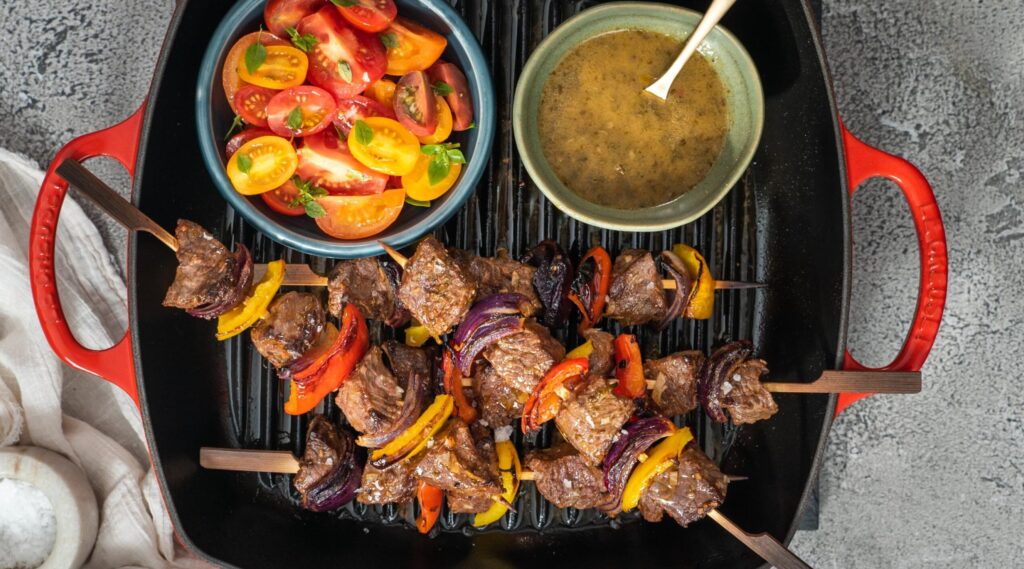 4 beef and vegetable skewers, a bowl of tomato salad and a bowl of greenish brown sauce on a red skillet with two handles.