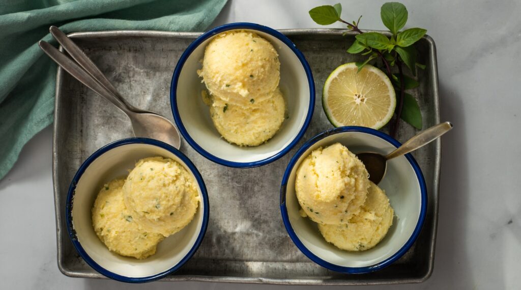 Three blue rimmed white bowls with two scoops of pale yellow ice cream in each, spoons, a cut lime half, herb on a metal tray on marble with green cloth.