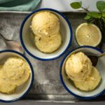 Three blue rimmed white bowls with two scoops of pale yellow ice cream in each, spoons, a cut lime half, herb on a metal tray on marble with green cloth.
