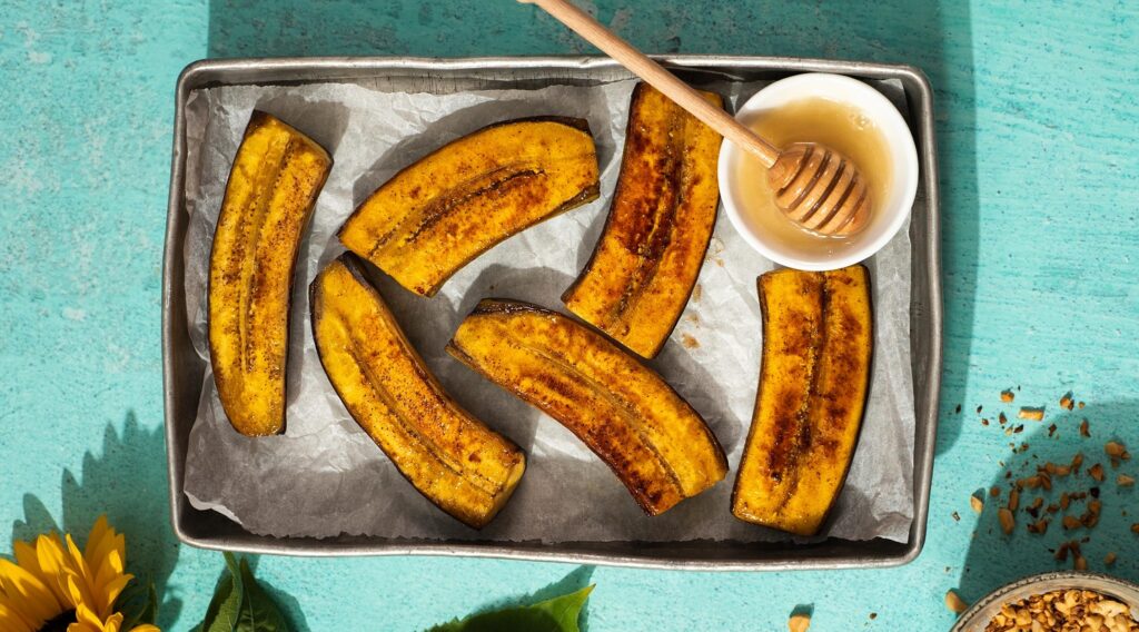 6 browned banana like food and a dish of honey with wooden stick on a metal tray on green mint green background. A sunflower and a bowl of nuts at the bottom edge.