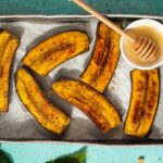 6 browned banana like food and a dish of honey with wooden stick on a metal tray on green mint green background. A sunflower and a bowl of nuts at the bottom edge.