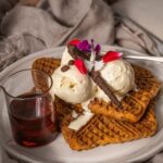 3 pieces of waffles topped with white ice-cream, chocolate and flower pettals and a jar of brown liquid on a white round plate on cloth with a cup of drink
